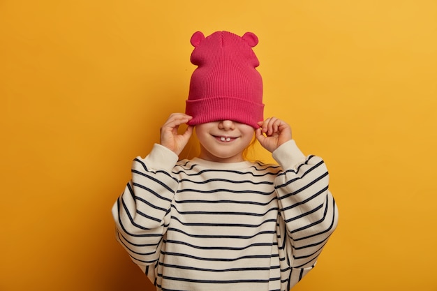 La foto de una niña alegre tiene dos dientes que sobresalen, cubre la mitad de la cara con un sombrero, usa un jersey de rayas casual, se divierte, se viste y se prepara para caminar al aire libre, aislado en una pared amarilla