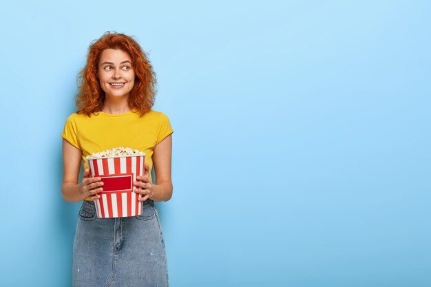 Foto de niña encantadora jengibre sostiene cubo con palomitas de maíz