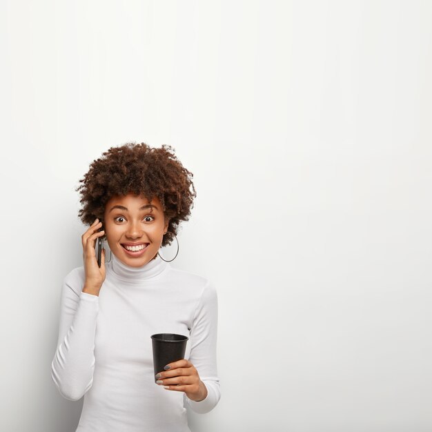 Foto de niña hipster de pelo rizado satisfecha recibe buenas noticias durante la conversación por teléfono móvil