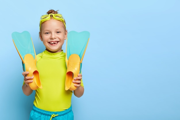 La foto de una niña pequeña de jengibre feliz muestra nuevas aletas de goma, usa gafas de natación, se viste con ropa de verano, disfruta aprendiendo a nadar, tiene descanso activo, aislado en una pared azul con espacio en blanco