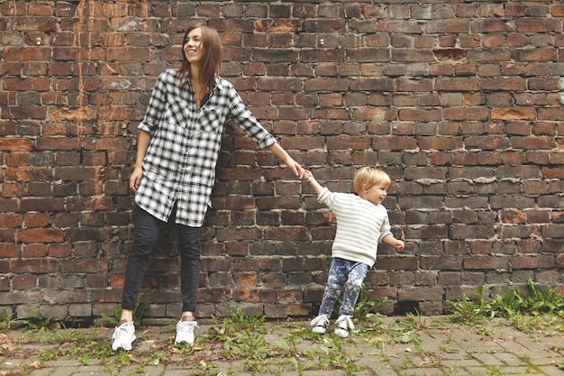 Foto de niño rubio y niña en un paseo cerca de la pared de ladrillo. Un tipo diminuto tirando de una tía adulta para que se vaya. Chica caucásica en camisa a cuadros permanece terca. Dos personas mirando a lados opuestos.