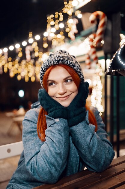 Foto nocturna al aire libre de una joven hermosa y feliz sonriente disfrutando de una decoración festiva, posando en la feria de Navidad, en la calle de la ciudad europea, usando un gorro de punto