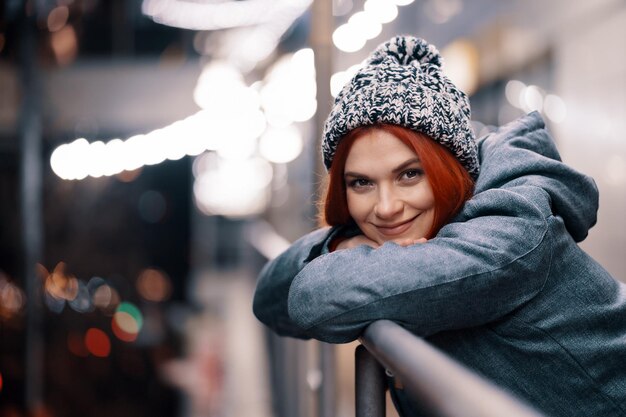 Foto nocturna al aire libre de una joven hermosa y sonriente feliz disfrutando de una decoración festiva, en la calle de una ciudad europea, usando un gorro de punto