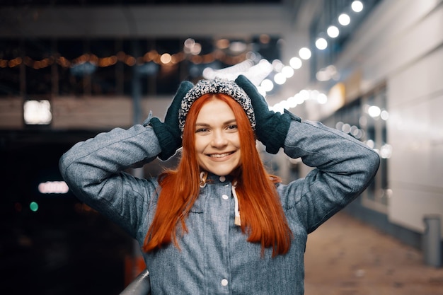 Foto gratuita foto nocturna al aire libre de una joven hermosa y sonriente feliz disfrutando de una decoración festiva, en la calle de una ciudad europea, usando un gorro de punto