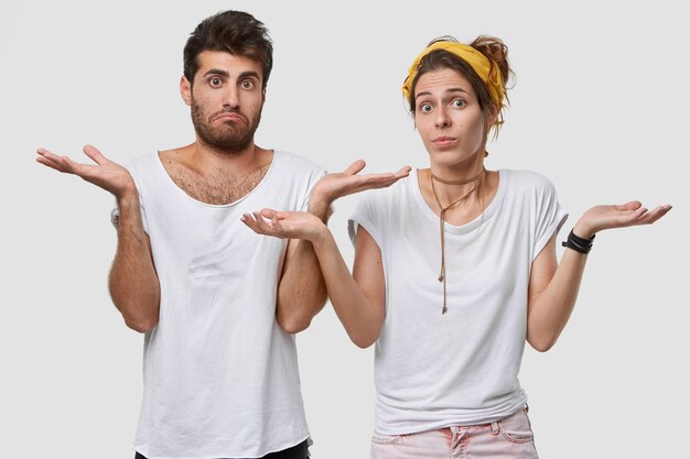 Foto de novios vacilantes que miran con incertidumbre, reciben sugerencias, tienen expresiones faciales dudosas, usan una camiseta blanca en un tono con la pared