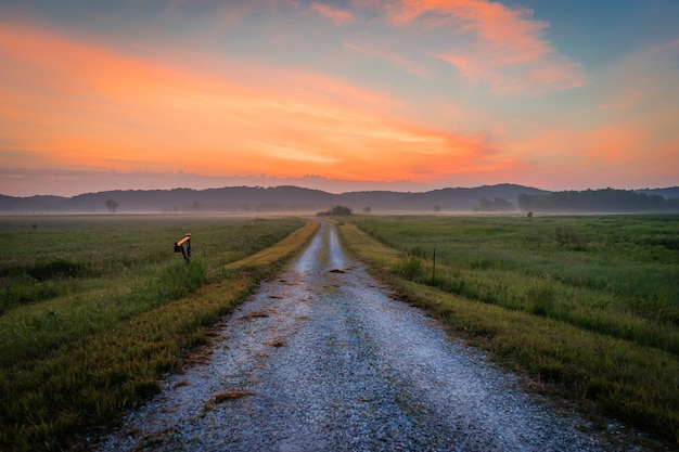 Foto panorámica de un camino de tierra en medio de un campo con la silueta de las montañas al atardecer