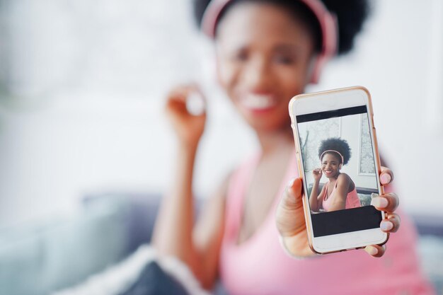 Foto de pantalla de teléfono móvil joven afroamericana sentada en el sofá escuchando música en los auriculares