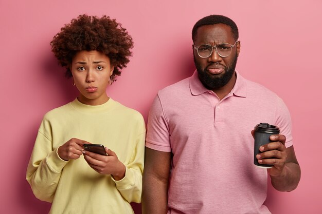 Foto de pareja afroamericana hosca desesperada que lee malas noticias en las redes sociales, bebe café para llevar, mujer triste señala en la pantalla del teléfono inteligente