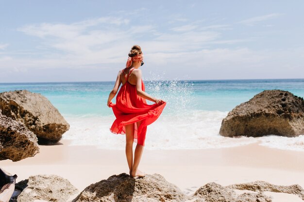 Foto de la parte de atrás de una chica bronceada bien formada de pie sobre una piedra grande. Disparo al aire libre de elegante modelo femenino jugando con su vestido rojo y mirando las olas del mar.