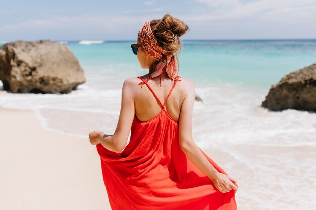 Foto de la parte de atrás de una hermosa mujer bronceada mirando al mar. Retrato al aire libre de increíble chica caucásica en vestido rojo de verano de pie.