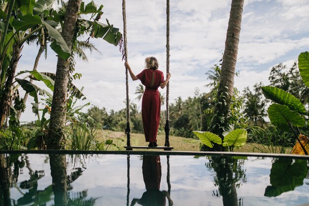 Foto de la parte posterior de la mujer delgada con vestido largo mirando el cielo lluvioso. Disparo al aire libre de modelo femenino bien proporcionado disfrutando de vistas a la naturaleza en el resort.
