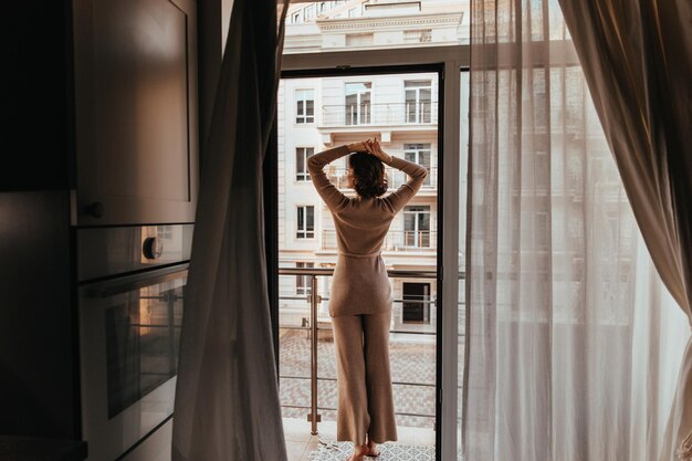 Foto de la parte posterior de una mujer con pantalones marrones Toma interior de una modelo caucásica mirando la ciudad a través de la ventana
