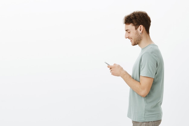 Foto de perfil del amable modelo masculino joven guapo en aretes, sosteniendo el teléfono inteligente en las manos, sonriendo ampliamente en la pantalla, satisfecho con el lindo mensaje de un amigo