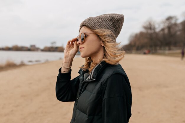 foto de perfil de cerca de la chica de moda con gorra y chaleco mirando al mar y tocando sus gafas de sol