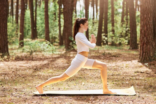 Foto de perfil completo de mujer adulta joven con vestidos de cola de caballo ropa deportiva elegante blanca haciendo yoga al aire libre, presionando las palmas juntas, meditación y relajación.