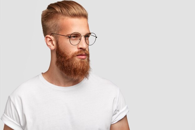 Foto de perfil de hombre brutal con barba espesa y astuta, usa gafas redondas y mira pensativamente a un lado