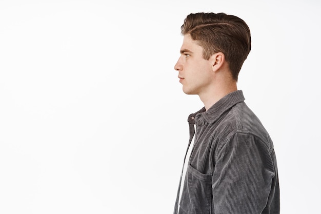 Foto de perfil de un joven serio, estudiante universitario adolescente mirando a la izquierda, mostrando un lado de la cara y el corte de pelo, de pie contra el fondo blanco