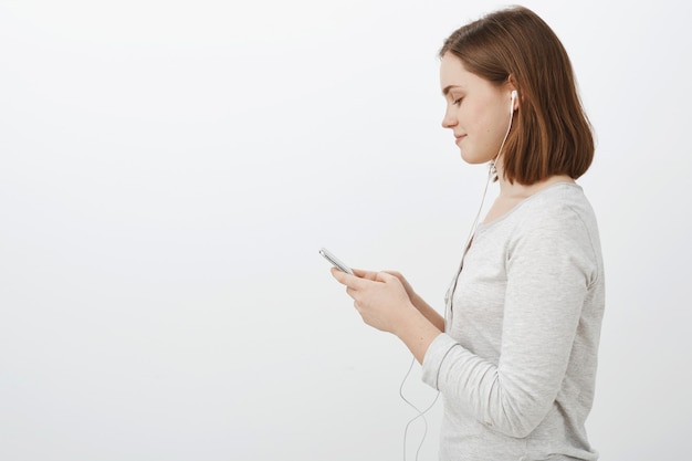 Foto de perfil de linda joven y sociable caucasain morena femenina con auriculares de pie contra la pared gris en una blusa blanca casual con smartphone sonriendo mientras envía mensajes de texto a un amigo en su camino a la fiesta