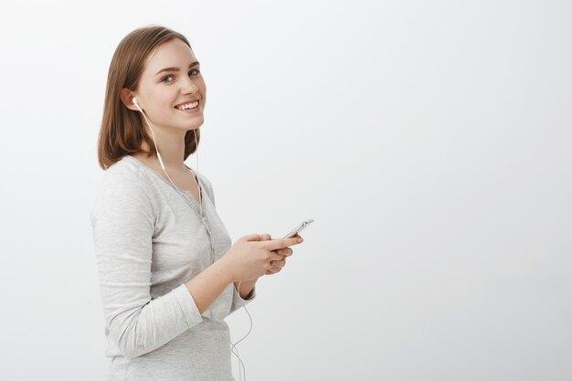 Foto de perfil de una mujer adulta despreocupada, guapa y entretenida con un corte de pelo corto y castaño que sostiene un teléfono inteligente mirando a un lado con una linda sonrisa satisfecha escuchando música en auriculares sobre una pared gris