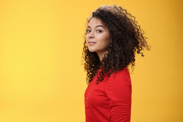 Foto de perfil de mujer europea de 25 años de pelo rizado atractiva elegante y fresca en vestido rojo girando a la cámara y sonriendo ampliamente con expresión despreocupada encantada, divirtiéndose sobre fondo amarillo.