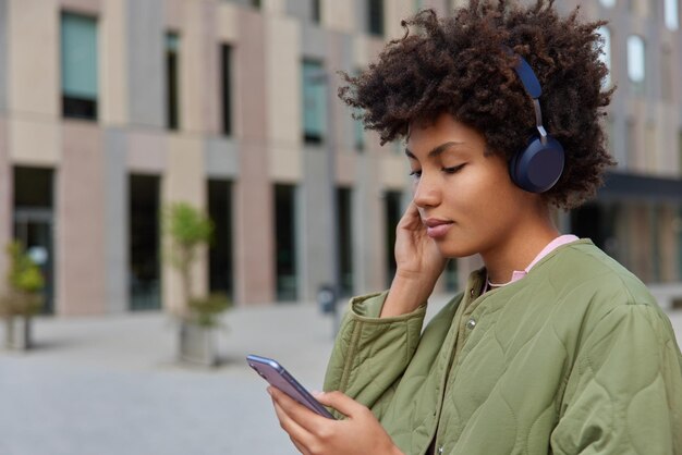 Foto de perfil de mujer de pelo rizado lee noticias a través del teléfono móvil lleva auriculares inalámbricos escucha la pista de audio de la lista de reproducción vestida con chaqueta