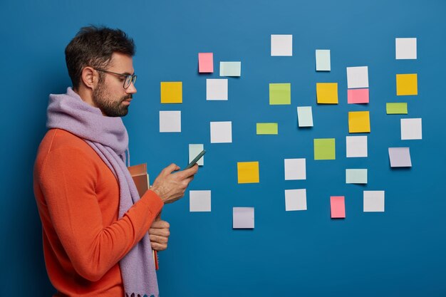 Foto de perfil de un profesional de negocios masculino vestido con ropa de abrigo, utiliza un teléfono móvil moderno para enviar mensajes de texto, sostiene libros de texto, pega papeles coloridos en el fondo