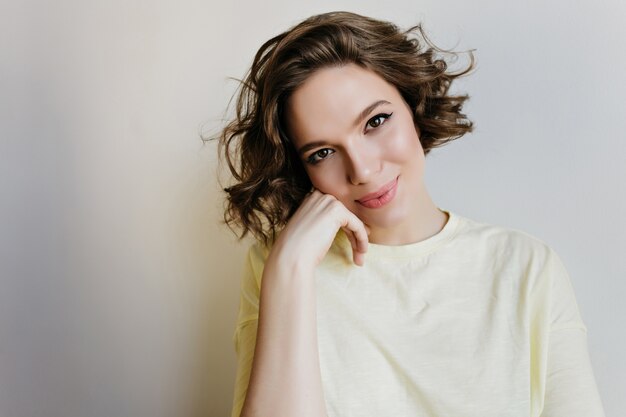 Foto de primer plano de adorable jovencita con sonrisa romántica. Retrato interior de una inspirada niña de pelo corto con hermosos ojos aislados en la pared blanca.