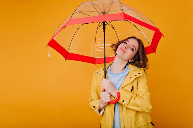 Foto de primer plano de adorable niña blanca sosteniendo paraguas y sonriendo suavemente. Retrato de interior de modelo femenino de pelo corto despreocupado en abrigo amarillo de otoño.