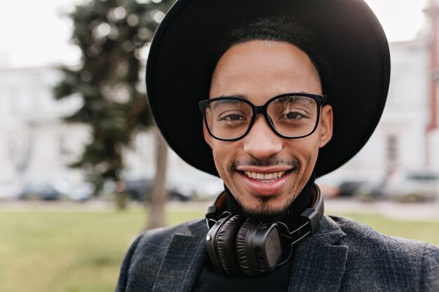 Foto de primer plano del feliz modelo masculino africano con ojos oscuros de pie sobre la naturaleza borrosa. Retrato al aire libre de chico elegante con sombrero negro y auriculares caminando por la calle.