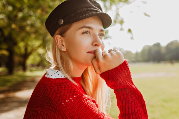 Foto en primer plano de una hermosa rubia disfrutando del sol en el parque de otoño. Hermoso retrato de mujer joven de pie bajo árboles amarillos.