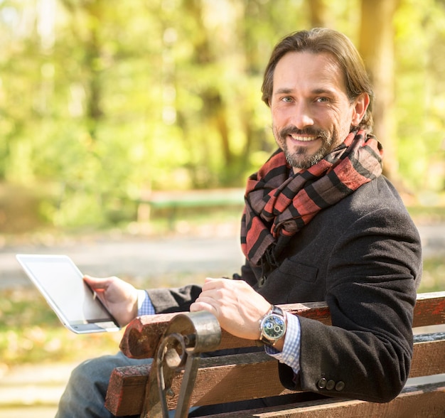 Foto gratuita foto de primer plano de un hombre guapo de mediana edad que trabaja en el parque hombre feliz con bufanda de moda sosteniendo una tableta frente a él al aire libre