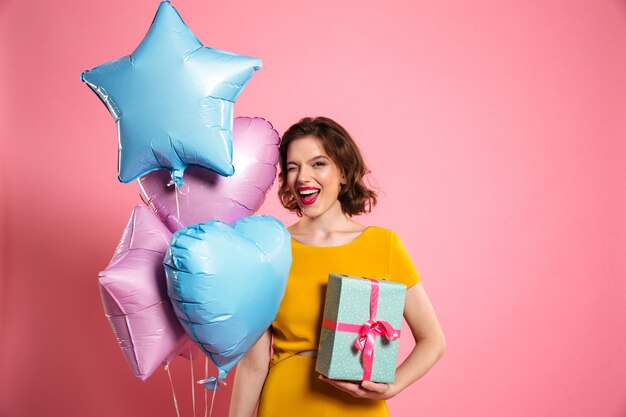 Foto de primer plano de juguetona cumpleañera con labios rojos guiña un ojo mientras sostiene globos y caja de regalo