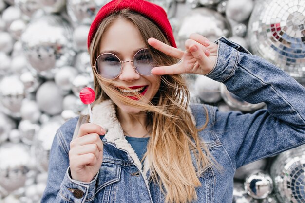 Foto de primer plano de magnífica chica rubia con expresión de cara feliz posando con bolas de discoteca. Retrato de señora guapa con sombrero rojo con piruleta en la pared urbana.