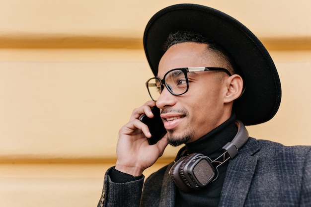 Foto de primer plano del modelo masculino americano con sombrero negro. Retrato al aire libre de apuesto hombre africano hablando por teléfono en la calle por la mañana.