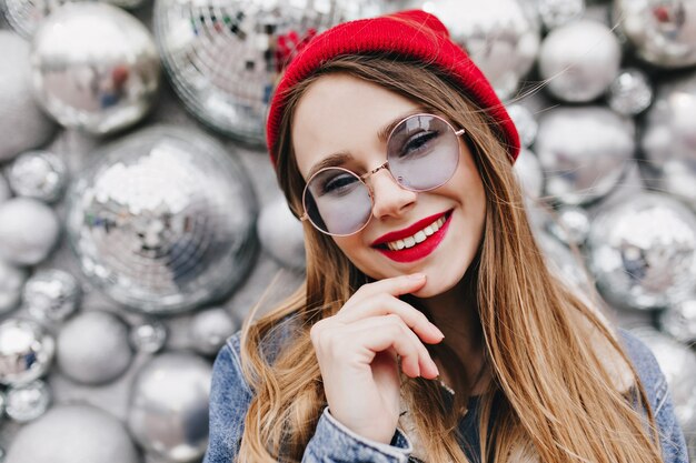 Foto de primer plano de niña complacida con maquillaje brillante posando cerca de bolas de discoteca. Retrato de mujer joven blanca con encanto en traje casual sonriendo