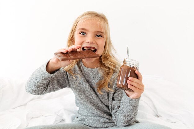Foto de primer plano de niña divertida comiendo barra de chocolate mirando a la cámara mientras está sentado en la cama
