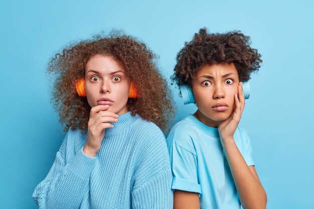 Foto de raza mixta dos mujeres de pie conmocionado mirada fija pose sin palabras de nuevo a eath otros tienen el pelo rizado llevar auriculares estéreo escuchar música aislada sobre la pared azul. Concepto de amistad