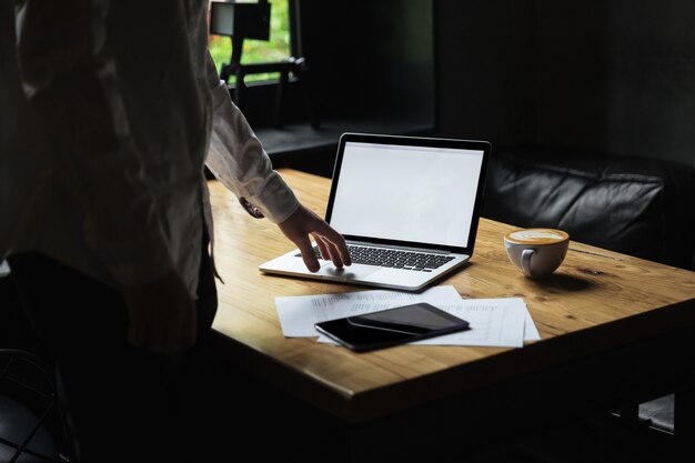 Foto recortada del empresario en camisa blanca de pie cerca de la mesa de madera, escribiendo en la computadora portátil