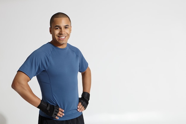 Foto recortada de feliz joven culturista afroamericano mirando a cámara y sonriendo con confianza, manteniendo las manos en la cintura, posando sobre fondo de pared de estudio blanco con espacio de copia para su texto