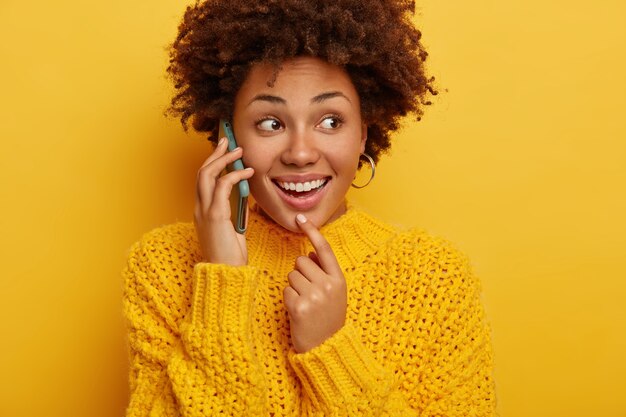 Foto recortada de feliz mujer afroamericana tiene conversación telefónica, sostiene el dispositivo cerca de la oreja, se centra a un lado, vestida con ropa de abrigo, aislado sobre fondo amarillo.