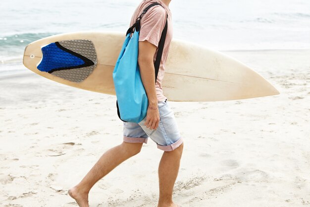 Foto recortada de un hombre descalzo con una bolsa azul que lleva una tabla de surf blanca en la mano, caminando por la costa arenosa mientras regresa a casa después de un entrenamiento activo de surf con otros surfistas