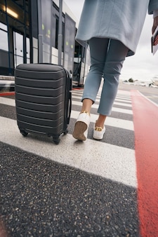 Foto recortada de una mujer con una maleta con ruedas y un boleto de embarque que se dirige hacia el autobús del aeropuerto