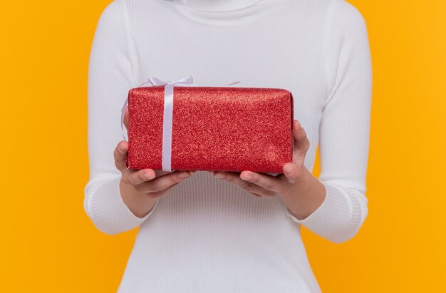 Foto recortada de mujer sosteniendo un regalo en manos de pie sobre la pared naranja