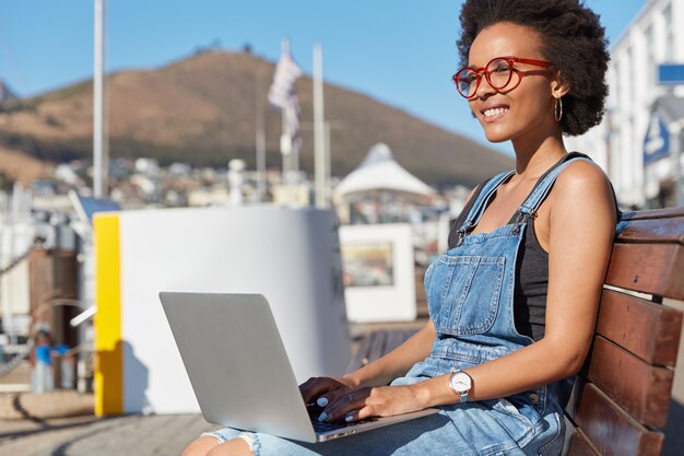 Foto de redactor publicitario o autónomo satisfecho escribe información sobre el teclado de una computadora portátil, piensa en algo creativo, usa ropa informal, modela en un banco al aire libre, trabaja como autónomo, usa 4G