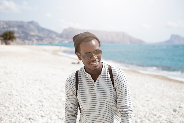 Foto de risa joven y guapo mochilero de piel oscura con lentes de espejo y sombrero sintiéndose feliz mientras caminaba por la playa durante el fin de semana de verano