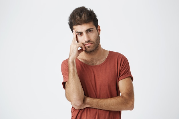 Foto gratuita foto de un serio y molesto joven estudiante bronceado sosteniendo su dedo en la sien y mirando con expresión concentrada, como si estuviera cansado de escuchar algo que había escuchado antes