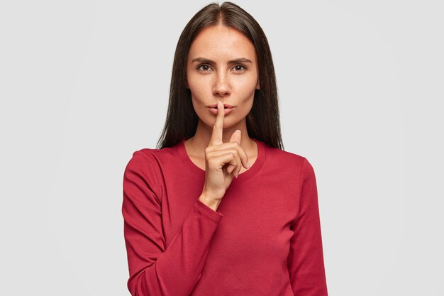 Foto de silenciosa hermosa dama morena vestida con suéter rojo, mantiene el dedo índice en los labios, tiene expresión seria,