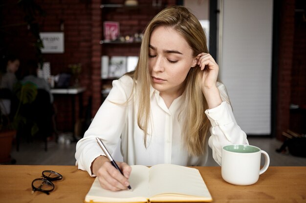 Foto sincera de una atractiva estudiante rubia en blusa blanca haciendo la tarea en el lugar de trabajo en casa, escribiendo en un cuaderno abierto, bebiendo té, con una expresión facial concentrada seria