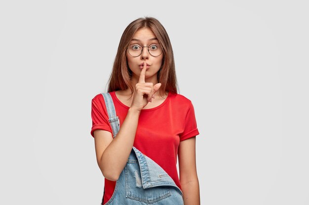 Foto sincera de una dama europea seria que hace un gesto de silencio, mantiene el dedo índice sobre los labios