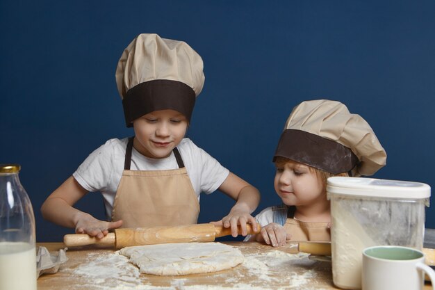 Foto sincera de la encantadora niña con gorro de cocinero mirando a su hermano mayor amasando masa para galletas o tarta